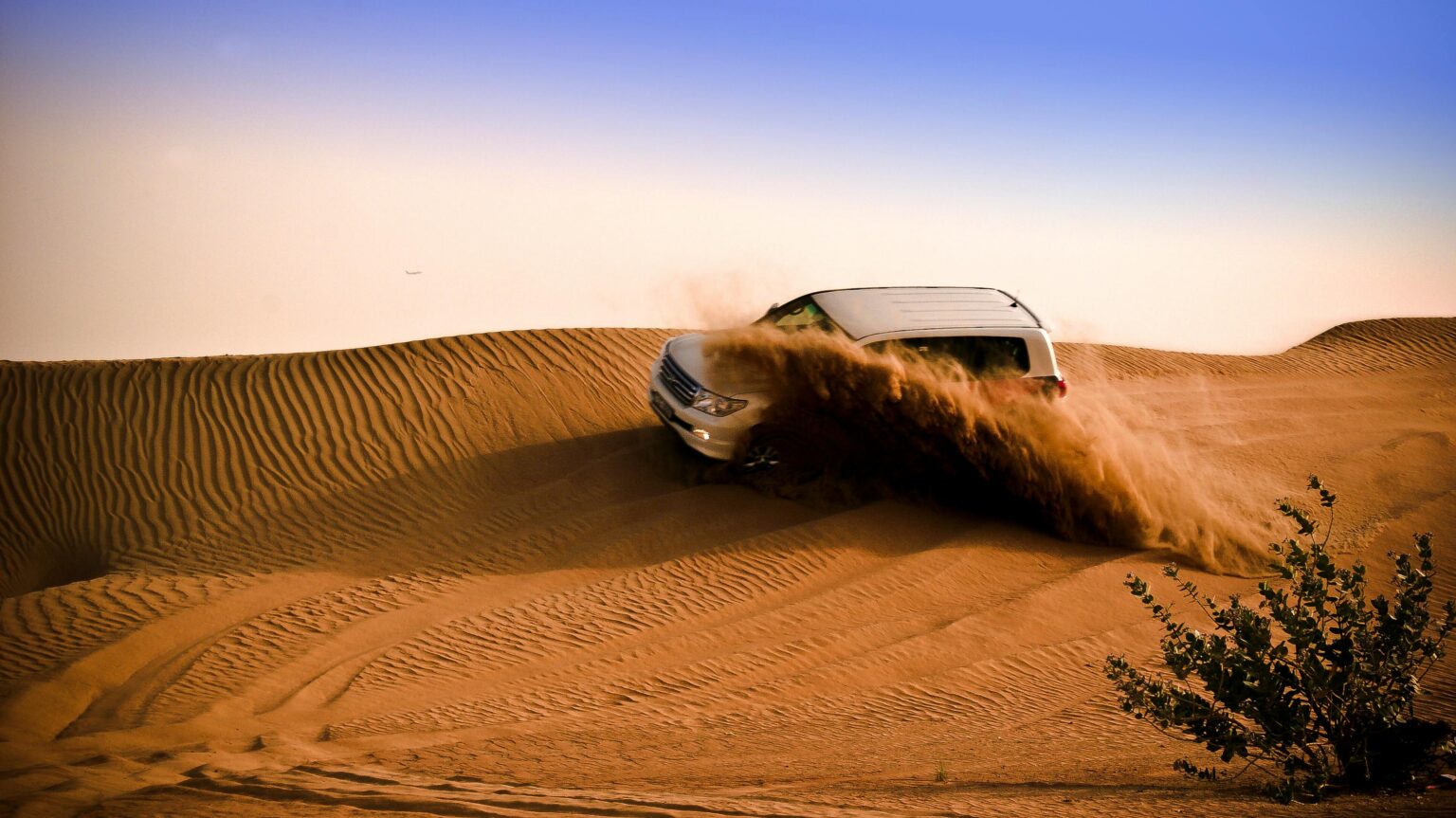 An SUV driving over sand dunes in the Dubai desert, with sand spraying as it moves